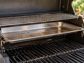 Stainless steel drip pan set inside a gas grill beneath the warming rack, catching drippings