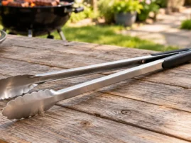 Long-handled stainless grill tongs lying on a weathered wooden table with a kettle grill behind